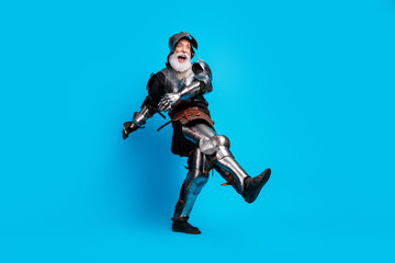 Senior man in medieval knight costume posing enthusiastically against blue background with a...