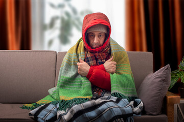 Middle age male model in hat parka and plaid getting cold sitting on the sofa in cold living room
