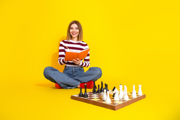 Young woman in casual style reading a book and playing chess on a vibrant yellow background