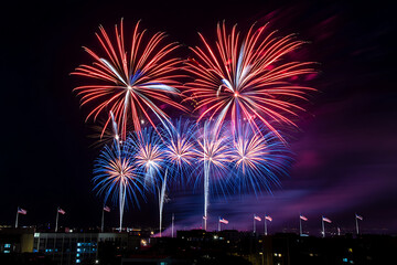 Obraz premium Brilliant red, white, and blue fireworks light up the night sky over a city with waving American flags in celebration of independence day