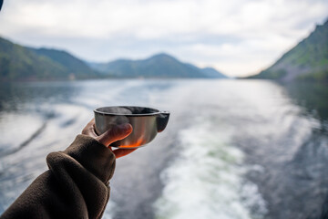 Hand holding hot drink in metal mug with view of Teletskoye lake in Altai Russia. Cozy moment in...