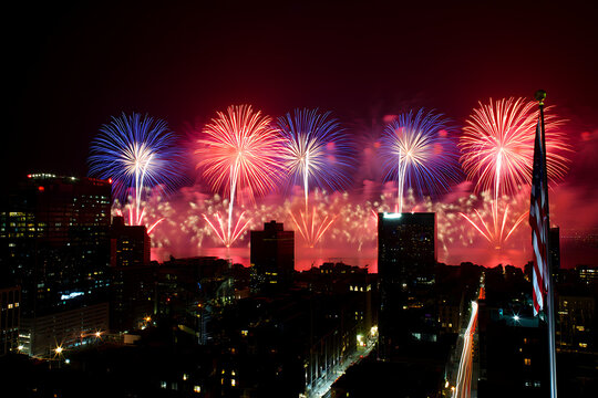 Spectacular fireworks illuminate the night sky over the city skyline with vibrant colors and the American flag waving in the foreground