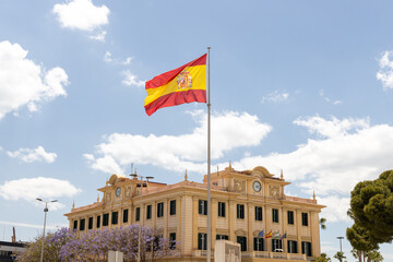 Port Authority of Malaga. National flag of Spain waving proudly in front of historic building under bright blue sky with fluffy white clouds creating a vibrant scene