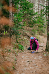 Obraz premium Woman Tying Shoe in Forest Trail.Female hiker crouching to tie her running shoe on a forest path surrounded by pine trees, dressed in sporty winter clothing.