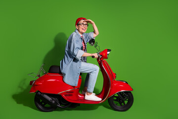 Middle aged woman enjoying a joyful ride on red scooter against vibrant green background in casual denim and stylish beanie