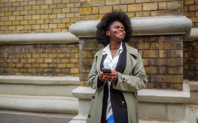 Young manager smiling while using her smartphone, leaning against a brick wall in the city. Enjoying break time, radiating positivity and confidence in a modern urban setting