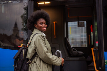 Smiling young black woman with afro hairstyle and backpack exiting a modern public transport bus while holding an umbrella, going to work or school