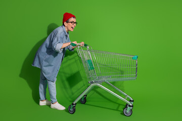 Smiling woman wearing trendy denim outfit and red beanie leaning on a shopping cart against a...