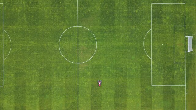 Aerial view of a red lawn mower cutting grass on a football field, creating striped patterns. Sports field maintenance in progress on a lush green surface