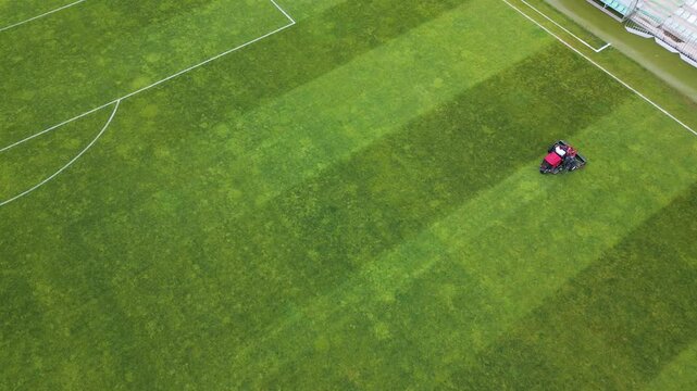 Aerial view of a red lawn mower cutting grass on a football field, creating striped patterns. Sports field maintenance in progress on a lush green surface