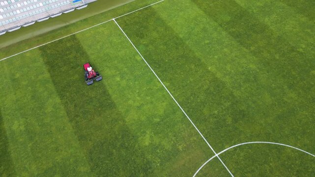 Aerial view of a red lawn mower cutting grass on a football field, creating striped patterns. Sports field maintenance in progress on a lush green surface