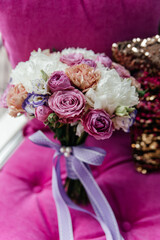 Wedding bouquet with pink roses, white peonies, and other flowers, tied with a light purple ribbon, rests on a fuchsia velvet chair, adding a touch of elegance and romance to the scene