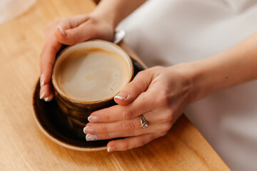 Woman with manicured nails and a diamond ring warming her hands on a cup of freshly brewed coffee, sitting at a wooden table, enjoying a quiet moment