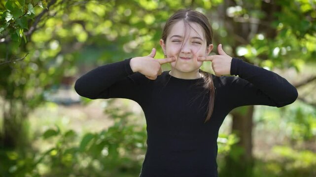 Teenage girl has fun in park, puffing out cheeks and pressing fingers on both sides to release air. Young female adolescent enjoys herself outdoors, inflating cheeks and playfully pushing on them to