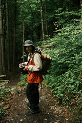Fototapeta premium Young man hiking through dense green forest with camera in hand, wearing backpack and rain jacket, exploring nature and documenting.