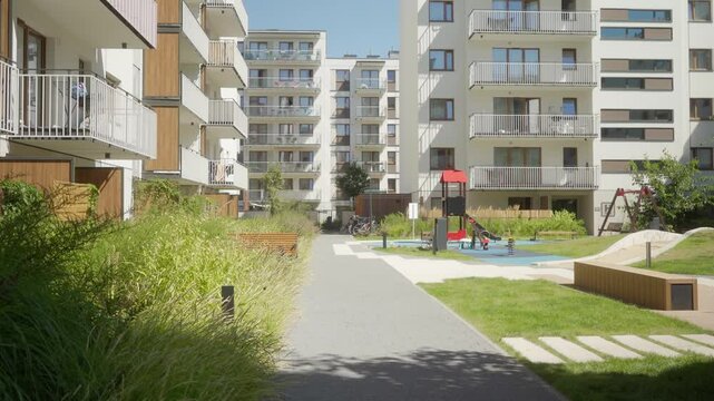 A modern residential complex of multi-family apartment buildings with a children&rsquo;s playground, landscaped recreational areas with benches, and pedestrian walkways within a gated area on a summer day.