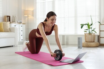 Happy woman exercising with ab wheel while watching video tutorial via laptop at home