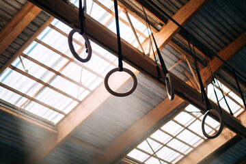 Fototapeta premium A rustic workshop or storage area with wooden beams and corrugated metal ceiling, illuminated by natural light Large hanging rings along a beam, varying positions casting shadows on th - AI-Generated