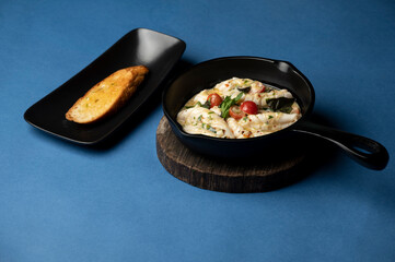 Pan-fried butter garlic baby squid served in a black pan on a wooden stand, accompanied by garlic bread in a black tray, styled on a bold blue background.
