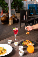 Hand picking a forkful of Sichuan chilli fish from a black plate, styled with colorful cocktails and pink sauce penne pasta on a table in vibrant restaurant setting.

