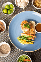 Top view of pan-seared chicken with vegetables and brown dip in a blue plate, surrounded by white bowls filled with lemon, raw garam masala, garlic, chili, and cumin on a table mat.