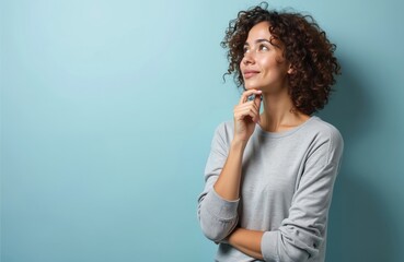 Young woman looks up thinking, smiling. Thoughtful expression on face, hand touches chin. Curly brown hair, neutral studio background. Pensive portrait, contemplation, decision-making.
