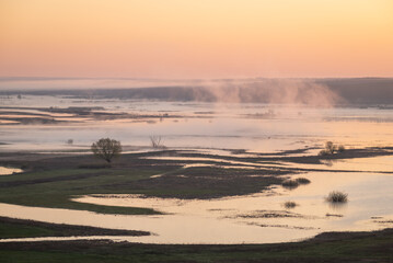 Morning fog envelops tranquil river landscape at dawn. Soft fog drifts over a serene flooded river landscape as dawn breaks, revealing calm waters and distant forests
