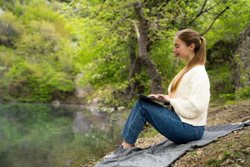 Full body side view of young woman in casual looking at laptop pc computer screen distance learn remote work rest relax in daylight sit near lake, freelance lifestyle, digital nomad outdoors in nature
