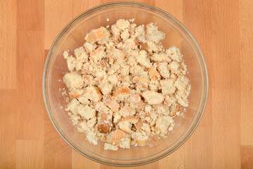 An overhead shot captures a glass bowl filled with moistened, crumbled stale bread, likely prepared for bread dumplings or stuffing, set on a wooden surface