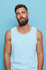 Obraz premium A young man with a well-groomed beard and short hair, wearing a light blue tank top mockup. He stands against a solid blue background, exuding a relaxed and confident demeanor.
