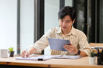 Young businessman analyzing data on digital tablet and writing notes at office desk.