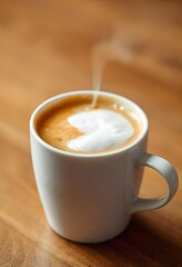 Obraz premium A white mug of hot coffee with a white foam heart design on top sits on a wooden table with steam rising from the drink