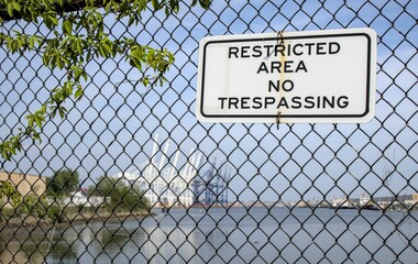 Naklejka premium restricted area no trespassing sign on black metal chain link fence in front of shipping docks in bayonne new jersey (infrastructure industrial ship container delivery security international tariff)