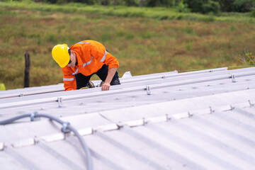 Worker in an orange safety uniform and helmet carefully inspecting a metal rooftop under natural...