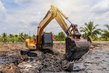 Excavator digging mud clay soil from pond bottom to expand and lift trench with level area for cultivation of garden furrows with water surrounding them in lowlands Thailand.
