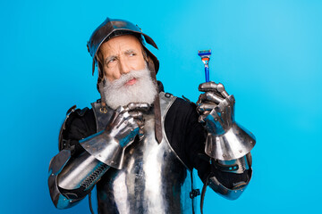 Portrait of a senior man dressed as a medieval knight holding a razor against a bright blue backdrop
