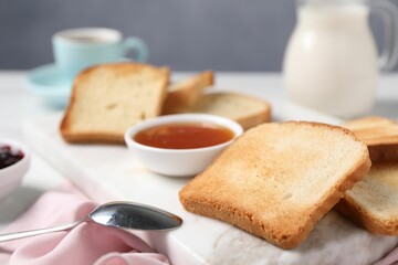 Slices of tasty toasted bread, spoon and jam on white table against grey background, closeup