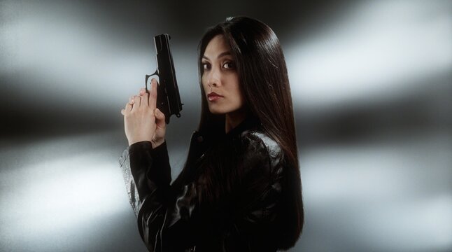 Asian woman holding a gun in a dark studio setting, wearing a black leather jacket