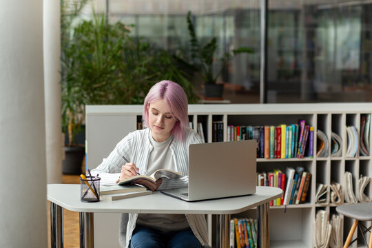 Attractive young female pink haired woman university student using a laptop while studying in modern libruary