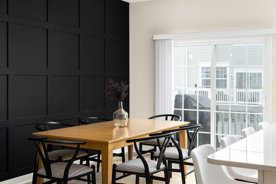 A cozy dining room with a wainscoting wall, wooden dining room table surrounded by black chairs, bar stools sitting at an island and a bright doorway.