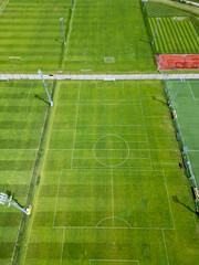 Aerial view of multiple football fields in a sports complex. Some have training lines, others standard markings. Green lawns, artificial turf, tennis court, and floodlights visible