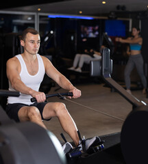 Sportive young man doing exercises on cable rowing device in well-equipped gym