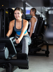 Fototapeta premium During workout, girl sits on simulator and pulls holder with her hands with effort. She does athletic exercises to give his muscles strength and endurance