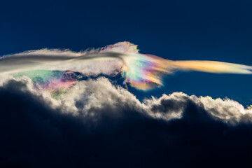 Spectacular iridescent cloud glows in vivid rainbow colors above dark storm clouds. Rare optical weather phenomenon captured against a deep blue sky. Amazing type of clouds.