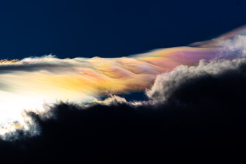 Spectacular iridescent cloud glows in vivid rainbow colors above dark storm clouds. Rare optical weather phenomenon captured against a deep blue sky. Amazing type of clouds.