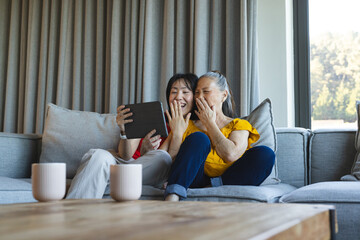 Laughing Asian mother and daughter leaning on sectional sofa in living room, with tablet and mugs