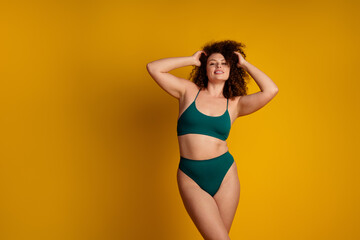 Confident plus size woman in green bikini against yellow background expressing natural beauty, acceptance, and charm
