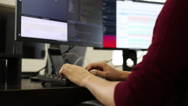 Programmer working at dual monitors, typing code in a dark-themed editor. Professional developer environment.