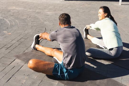 Diverse couple stretching on sandy beach using exercise mats, one smiling and one focusing