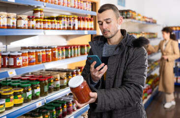 Adult man shopper with mobile phone chooses lecho in grocery store